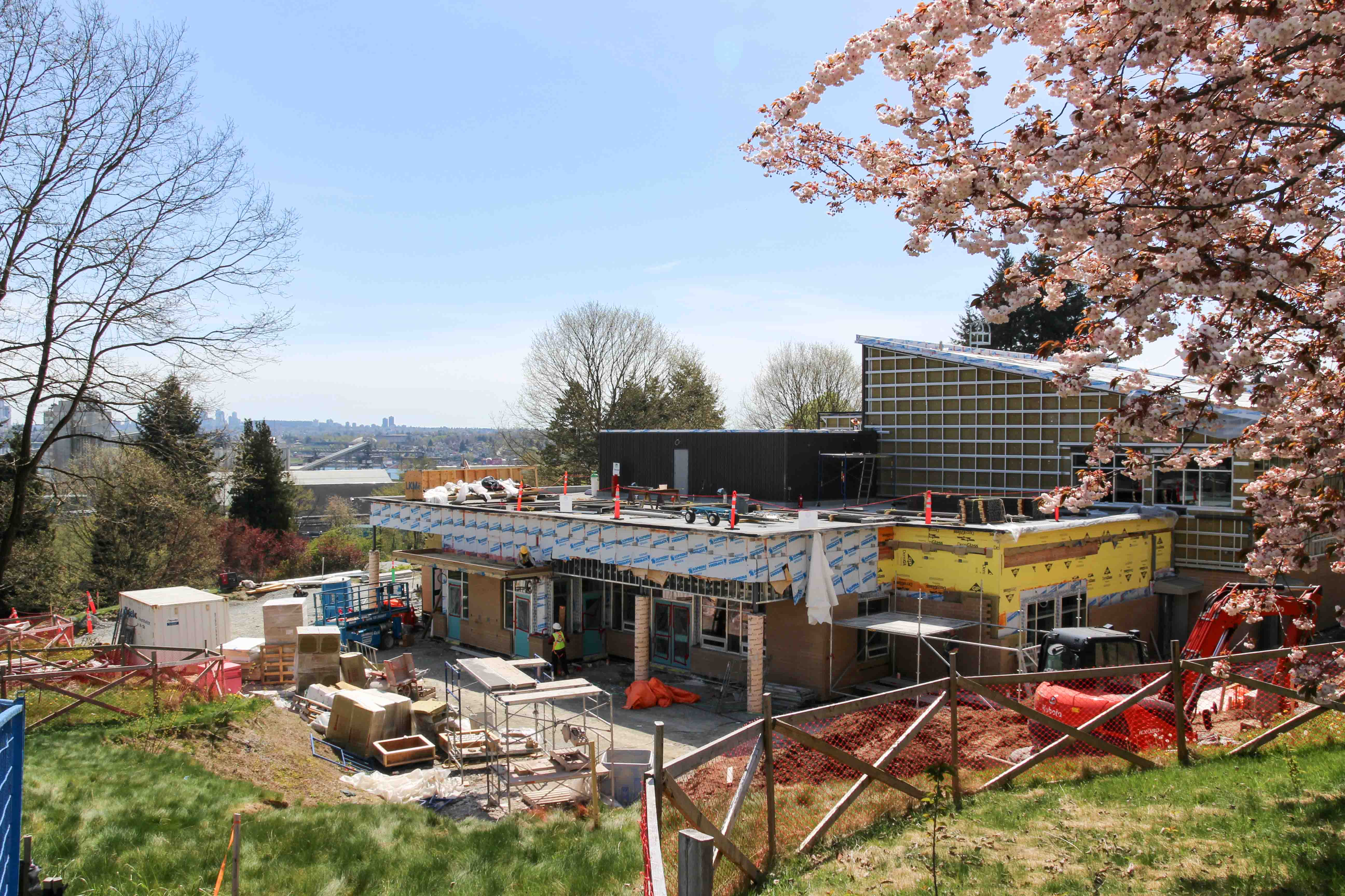 Outdoor view of a building under construction on a hillside, surrounded by trees and spring blossoms. Workers, scaffolding, materials, and equipment fill the site, with a city skyline and bridge visible in the distance under a clear blue sky.