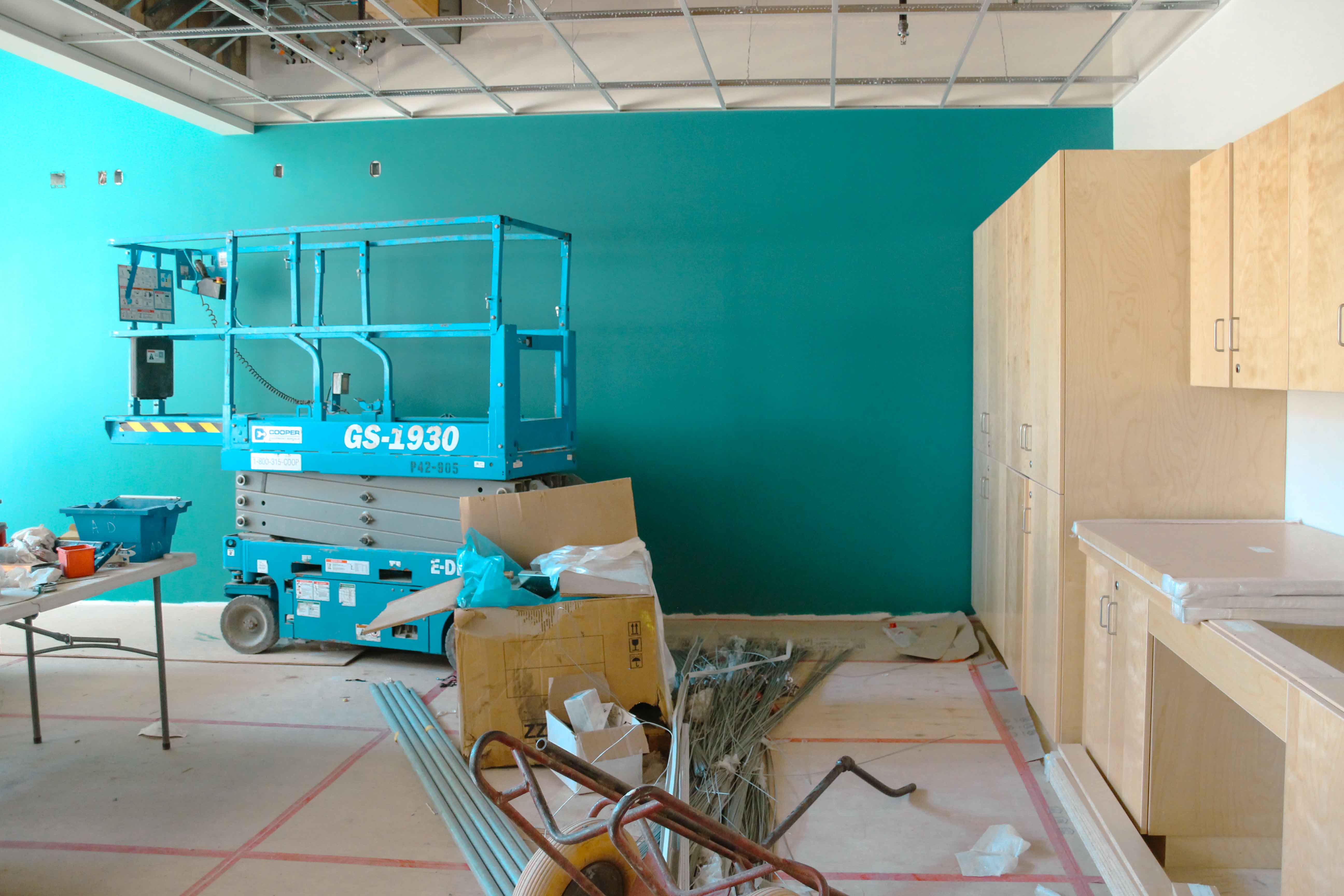 Indoor construction scene with a bright teal wall, exposed ceiling grid, and a blue scissor lift labeled “GS-1930.” Light wood cabinets line the right wall, while the floor is covered with paper, tools, pipes, and scattered building materials.