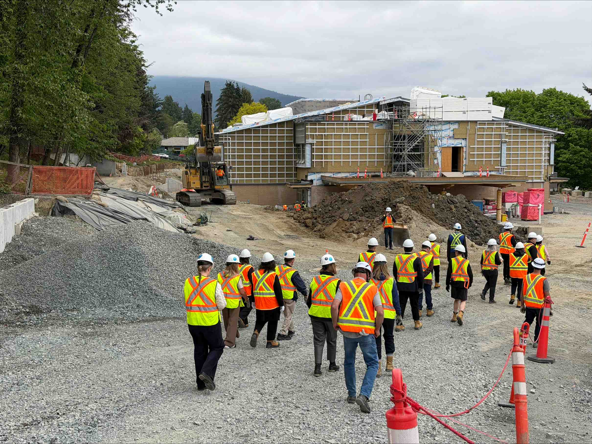 Construction site with a group of people wearing high-visibility safety vests and white hard hats walking along a gravel path. An excavator, soil piles, scaffolding, and an unfinished building are visible, with trees and mountains in the background under an overcast sky.