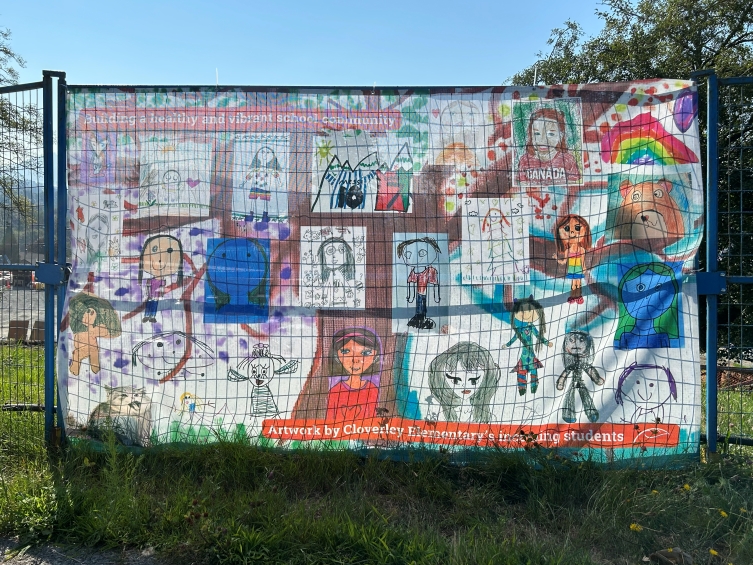 Large outdoor banner on a school fence featuring a colourful collage of student artwork, including portraits, landscapes, animals, a rainbow, and a globe. Text reads “Building a healthy and vibrant school community” and credits Cloverley Elementary students.