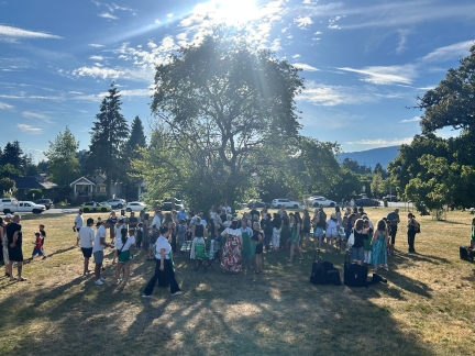 Large group of people gathered outdoors in a grassy park for an ice cream social. The crowd stands beneath a large tree on a sunny day, with long shadows stretching across the dry grass. Houses, parked cars, and forested mountains are visible in the background under a bright blue sky.