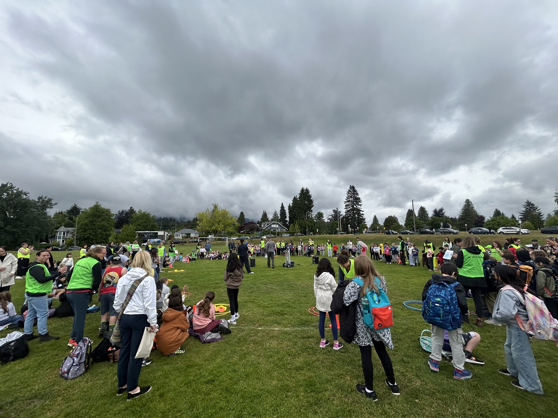 Large group of children and adults gathered on a grassy field, standing and sitting in a wide circle during an outdoor school event. Many wear bright green vests or backpacks. Houses, trees, and parked cars line the background beneath a cloudy sky.